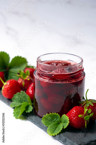 Strawberry jam in glass jar on slate dish with fresh strawberry fruit and green leaves on marble. Recipe of delicious berry jam of strawberry full of vitamins and antioxidants.
