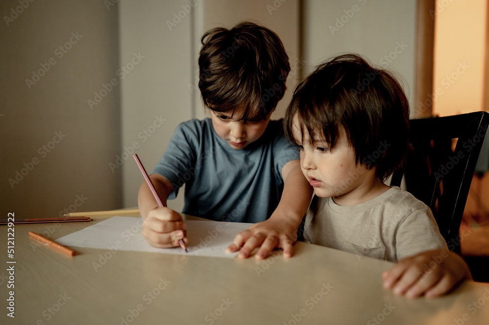 two brothers sit at home at the table and draw with colored pencils, the older brother teaches the younger one to draw, the concept of family and friendship two brothers sit at home at the table and draw with colored pencils, the older brother teaches the younger one to draw, the concept of family and friendship