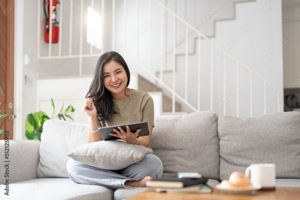 Beautiful attractive young Asian woman sitting on gray sofa and using digital tablet at living room in the morning. Beautiful attractive young Asian woman sitting on gray sofa and using digital tablet at living room in the morning.