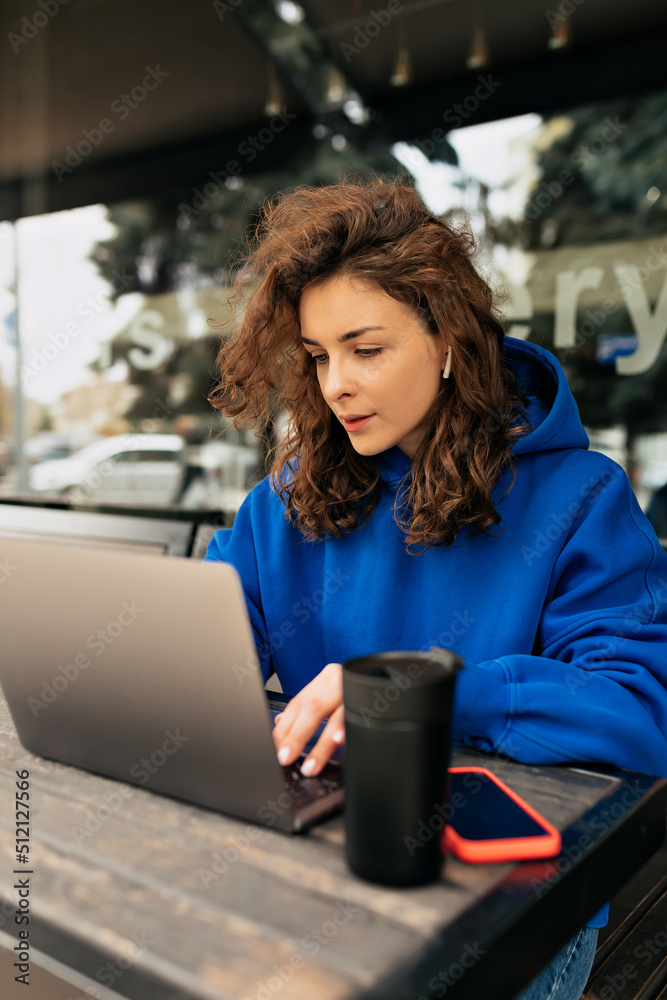 Focused caucasian woman working with computer at table outdoor in spring day. Brunette looks displeased at notebook screen. Freelance work, business people concept