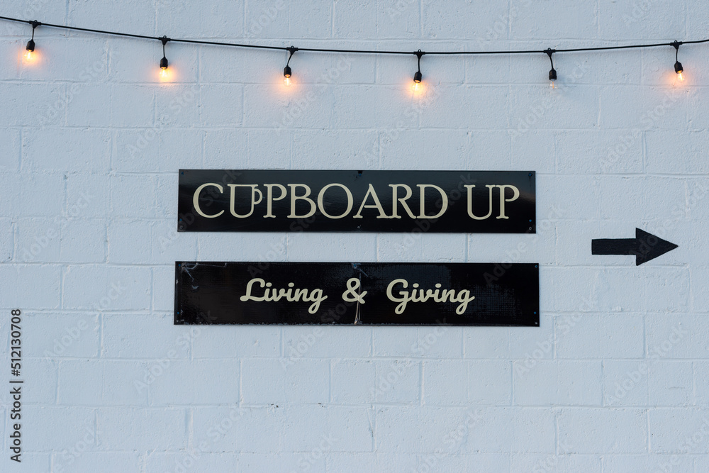 A white brick wall with a string of clear illuminated lights hanging from the top of the wall. There are two signs on black backing with white lettering. The sign's text is cupboard up, living  giving