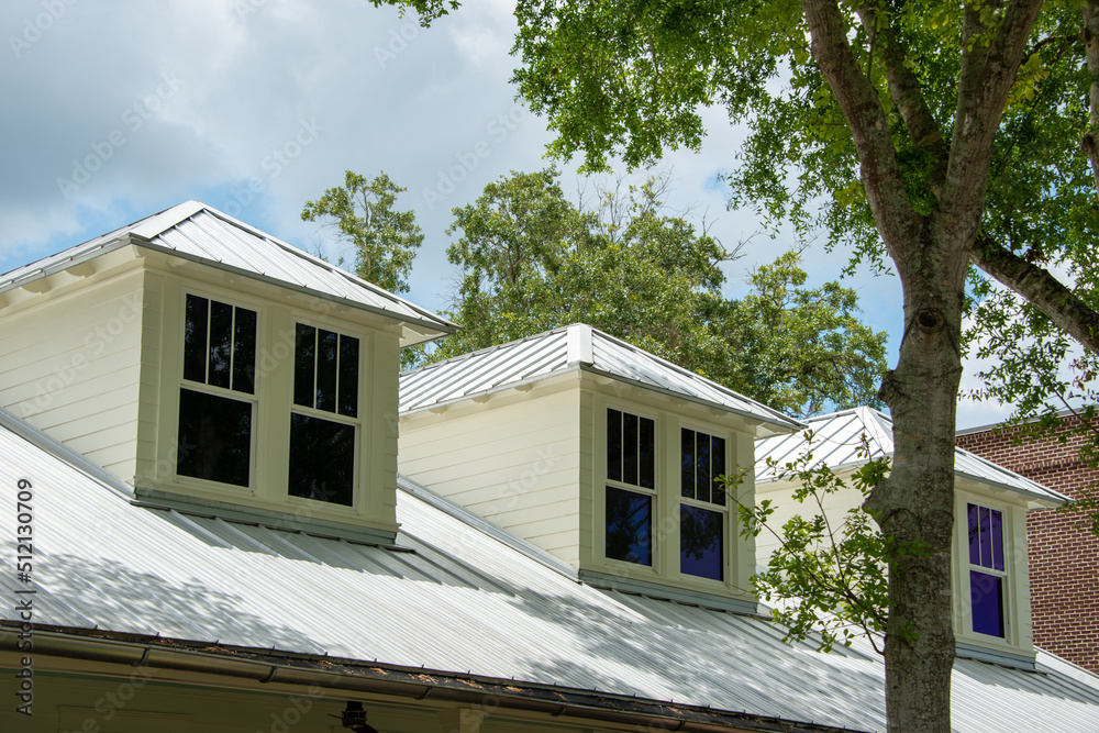 Three dormers with multiple double hung windows, a grey metal roof, and pale yellow vinyl siding on a large house. There are tall trees with thick green leaves both in front and behind the building. Three dormers with multiple double hung windows, a grey metal roof, and pale yellow vinyl siding on a large house. There are tall trees with thick green leaves both in front and behind the building.