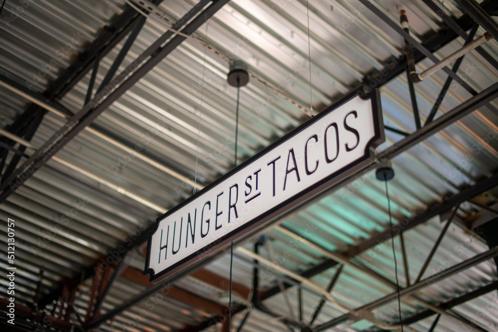 A white with black lettering sign hanging from the ceiling of a market. The sign spells hunger street tacos. The interior of the industrial warehouse ceiling is silver metal with a black grid system. A white with black lettering sign hanging from the ceiling of a market. The sign spells hunger street tacos. The interior of the industrial warehouse ceiling is silver metal with a black grid system.