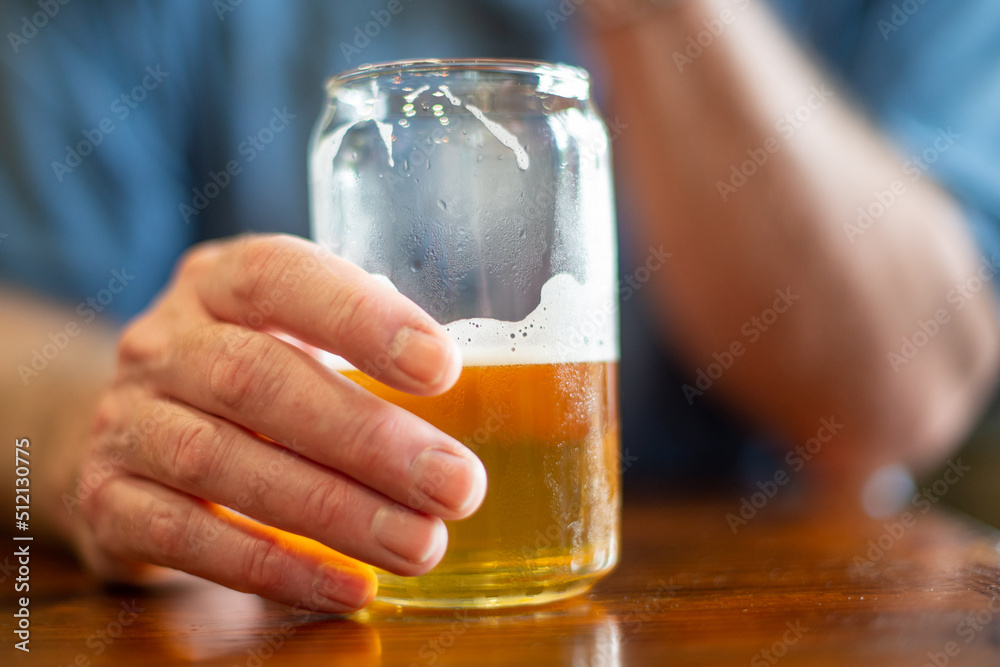 A male reaches for a tall clear glass of sour craft beer in a micro brewery. The cold refreshment has frost on the glass. The beer is on a table and coaster. The top of the glass has white froth. A male reaches for a tall clear glass of sour craft beer in a micro brewery. The cold refreshment has frost on the glass. The beer is on a table and coaster. The top of the glass has white froth.