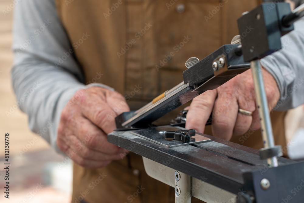 A man using a fixed angle knife sharpener kitchen tools at a farmer's market to sharpen knives for customers. The stainless steel kit has a long narrow holder for water stones of various grits. 