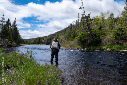 A male salmon angler stands in a river up to his waist with a long fishing rod wearing waders, a fly hat, and a vest. The background is trees and a riverbank. The blue water has a small ripple on top.