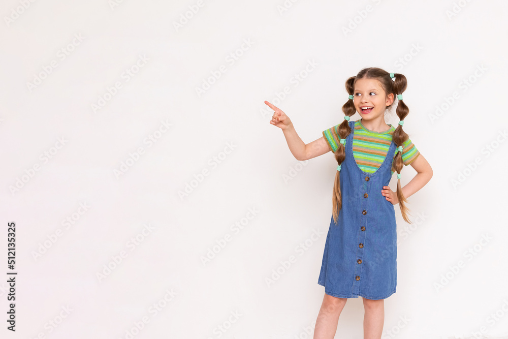 The child points sideways with his index finger at your advertisement on a white isolated background. A beautiful girl in a denim sundress. Copy Space The child points sideways with his index finger at your advertisement on a white isolated background. A beautiful girl in a denim sundress. Copy Space