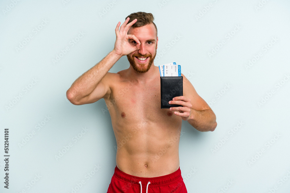 Young caucasian man holding a passport isolated on blue background excited keeping ok gesture on eye. Young caucasian man holding a passport isolated on blue background excited keeping ok gesture on eye.