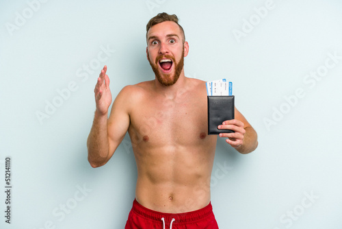 Young caucasian man holding a passport isolated on blue background receiving a pleasant surprise, excited and raising hands.