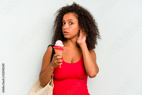 Young brazilian woman going to the beach holding an ice cream isolated on blue background trying to listening a gossip.