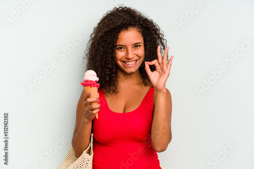 Young brazilian woman going to the beach holding an ice cream isolated on blue background cheerful and confident showing ok gesture.