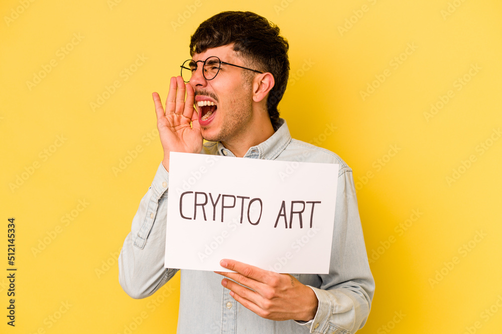 Young hispanic man holding crypto art placard isolated on yellow background shouting and holding palm near opened mouth. Young hispanic man holding crypto art placard isolated on yellow background shouting and holding palm near opened mouth.