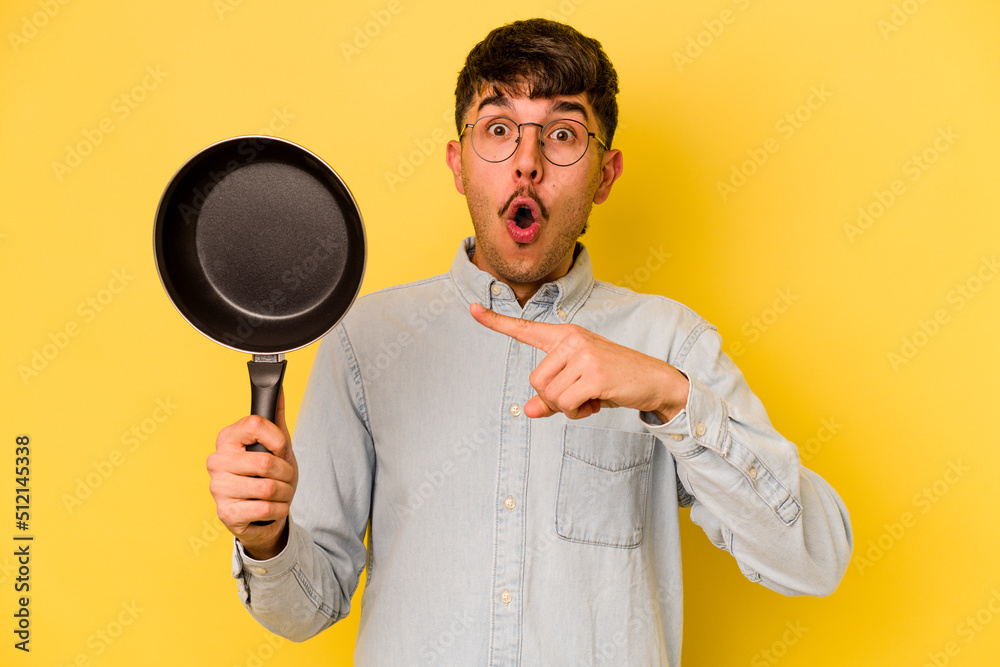 Young hispanic cooker holding frying pan isolated on yellow background pointing to the side Young hispanic cooker holding frying pan isolated on yellow background pointing to the side