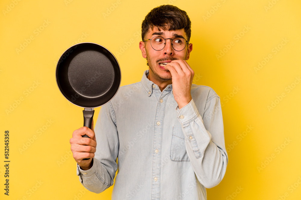 Young hispanic cooker holding frying pan isolated on yellow background biting fingernails, nervous and very anxious. Young hispanic cooker holding frying pan isolated on yellow background biting fingernails, nervous and very anxious.