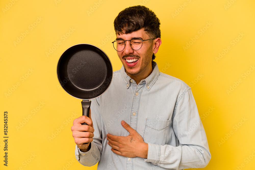 Young hispanic cooker holding frying pan isolated on yellow background laughing and having fun. Young hispanic cooker holding frying pan isolated on yellow background laughing and having fun.