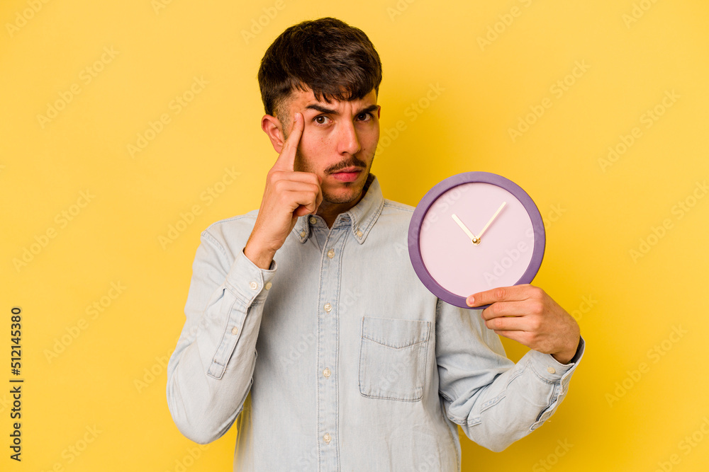 Young hispanic man holding a clock isolated on yellow background pointing temple with finger, thinking, focused on a task. Young hispanic man holding a clock isolated on yellow background pointing temple with finger, thinking, focused on a task.