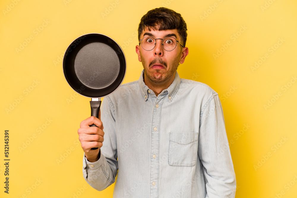 Young hispanic cooker holding frying pan isolated on yellow background shrugs shoulders and open eyes confused. Young hispanic cooker holding frying pan isolated on yellow background shrugs shoulders and open eyes confused.