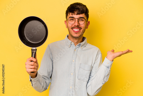 Young hispanic cooker holding frying pan isolated on yellow background showing a copy space on a palm and holding another hand on waist.