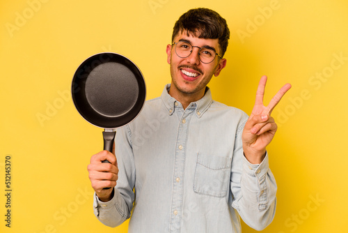 Young hispanic cooker holding frying pan isolated on yellow background joyful and carefree showing a peace symbol with fingers.