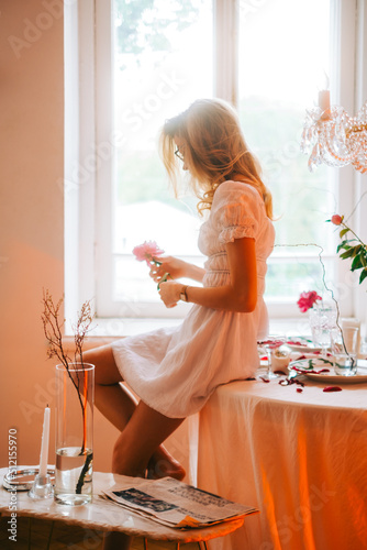 Portrait of attractive caucasian woman in a white dress sitting near window and holding flower in a hand.