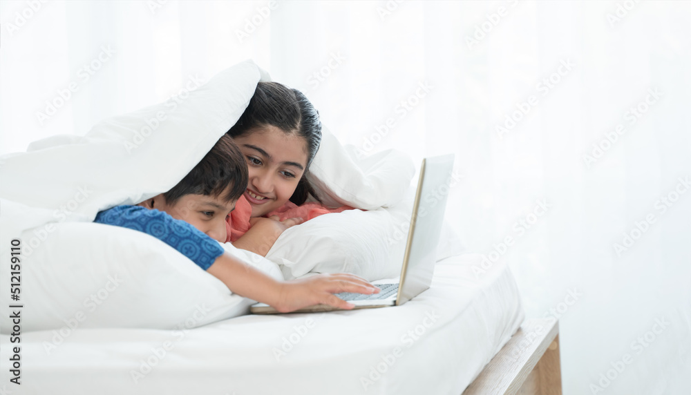 Two happy Indian brother and sister in traditional clothing lying on bed under blanket smiling, using laptop, having fun together at home. Education, Siblings relationship concept. Selective focus Two happy Indian brother and sister in traditional clothing lying on bed under blanket smiling, using laptop, having fun together at home. Education, Siblings relationship concept. Selective focus