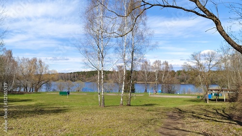 On the shore of the lake grow birches and other trees, there are cabins for changing clothes and created sports fields. There is a forest on the opposite shore. It is sunny, the sky is blue with cloud