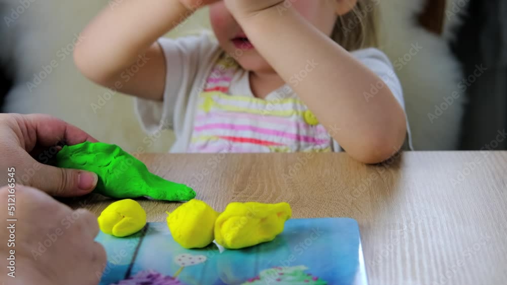 close-up plasticine toys in hands of toddler, small child, blonde girl 2 years old sculpts figures of characters from colored dough, concept of fine motor skills, tactile sensations, creativity