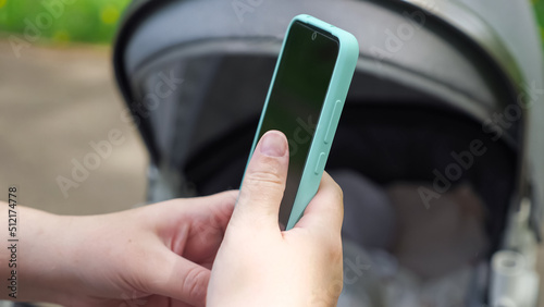 Adult hands holding phone scroll internet at baby stroller on sunny day. Mother relaxes with sleeping little daughter in park on blurred background closeup
