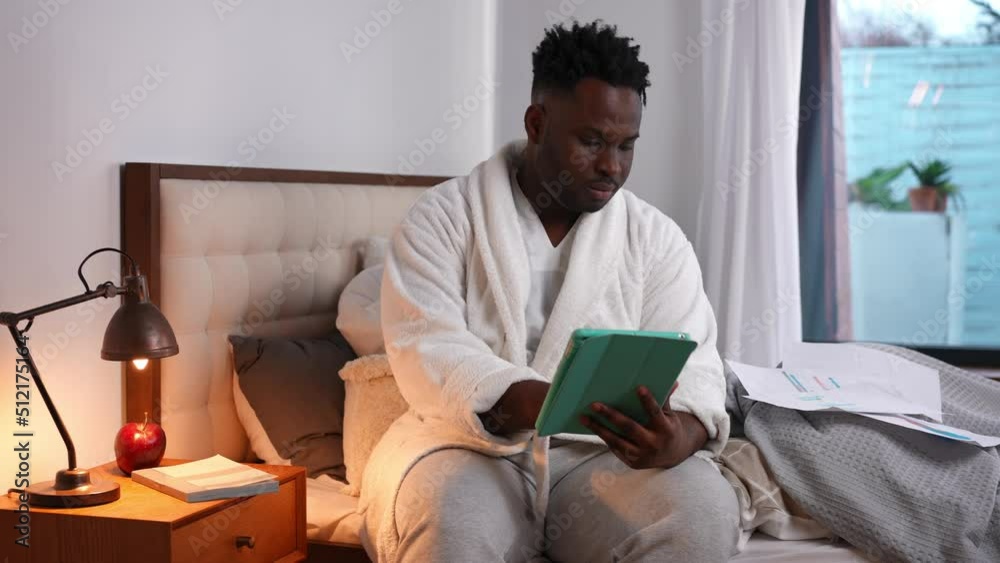 Smart handsome African American man analyzing e-market and documents sitting on bed at home. Portrait of focused intelligent young freelancer working online indoors. Lifestyle and e-business Smart handsome African American man analyzing e-market and documents sitting on bed at home. Portrait of focused intelligent young freelancer working online indoors. Lifestyle and e-business