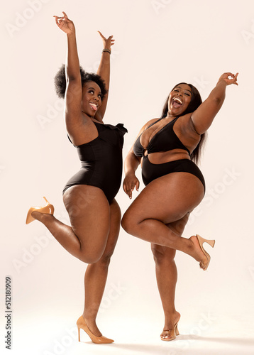 Full photo of superb plus size dark skinned two women in black fashionable swimwears, laughing and dancing, having fun together. Concept of body acceptance, body positivity photo