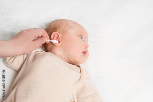 Mother tries to clean wax from ear of newborn baby using cotton swab. Adorable little girl lies on bed obstructing hygiene procedure close-up photo