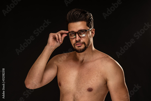 Handsome sensual man with brown eyes wearing eyeglasses, posing shirtless on black studio background. Muscular body.