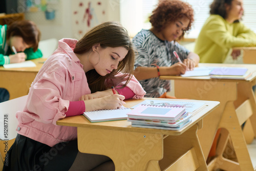 Adolescent girl in casualwear making notes in copybook while sitting by desk against her intercultural classmates at lesson of biology