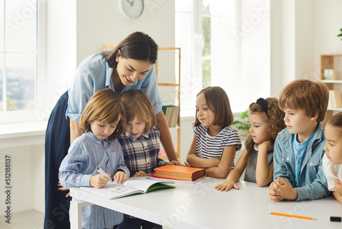 Young school teacher helping her happy elementary students who are writing in notebooks. Little children learning new things in class. Group of talented kids watching their classmate draw a picture