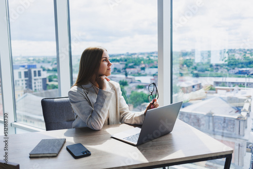 A female head of a company in glasses sits at a laptop in her office with a breathtaking view of the city. Business lady works at a wooden table.