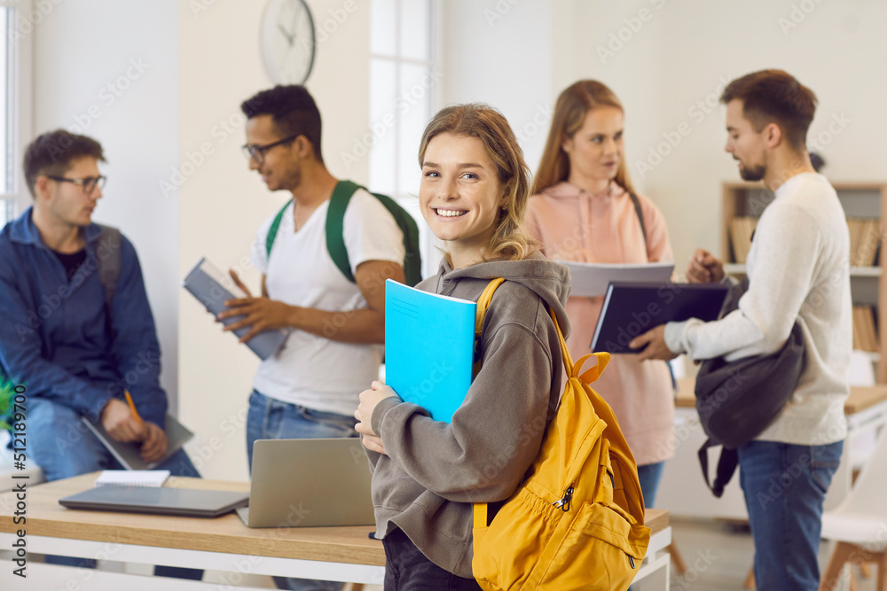 Portrait of happy smiling beautiful female university student with bag and notebook. Pretty college girl with candid cheerful face expression standing in classroom with her classmates in background - obrazy, fototapety, plakaty 