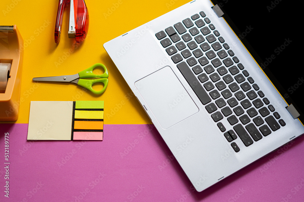 Office supplies, next to a laptop, on a yellow and pink split background. Concept of back to work in the office. Office supplies, next to a laptop, on a yellow and pink split background. Concept of back to work in the office.
