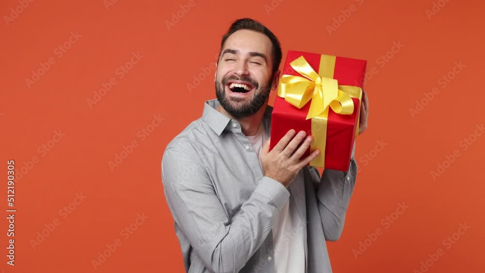 Excited jubilant exultant young brunet man 20s years old wears blue shirt hold shake red present box with gift ribbon bow try to guess what inside isolated on plain orange background studio portrait