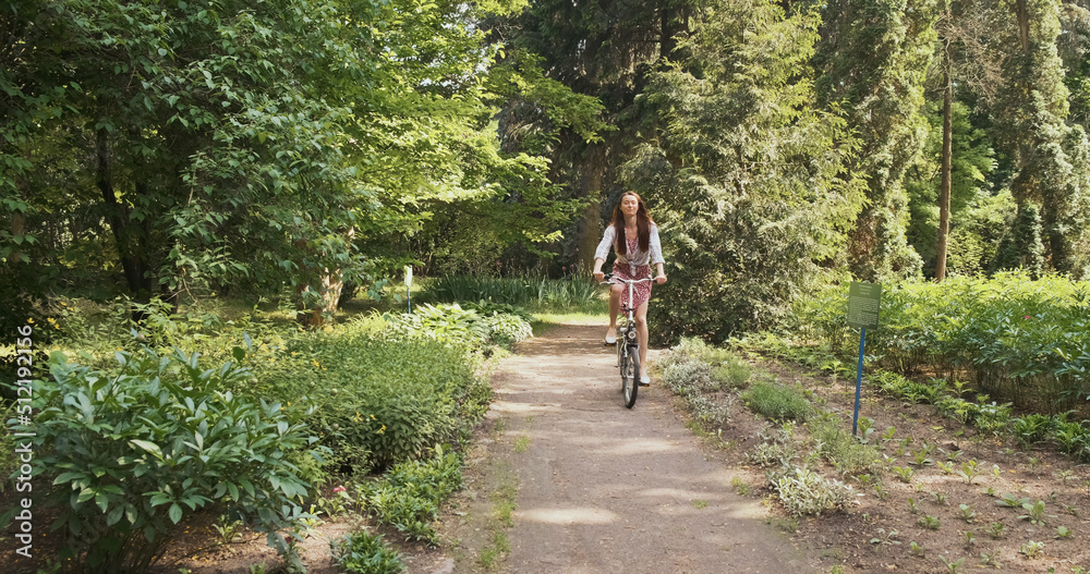 Fototapeta premium young woman rides a folding bike through a park with green trees towards the camera. The concept of a healthy lifestyle, eco movement, green transport. Wide shot, spring summer, daytime