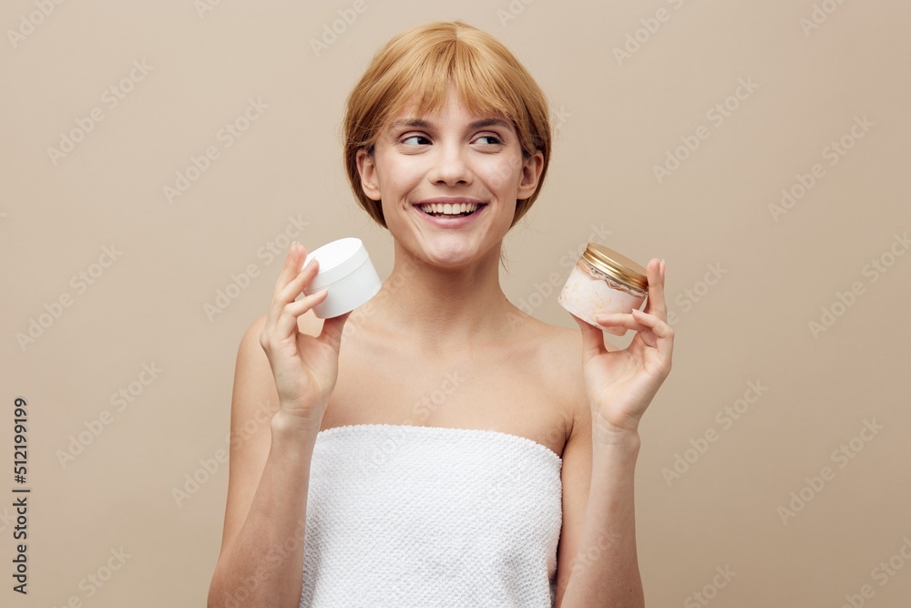 A joyful woman in a towel with beautiful skin, a beautiful smile and red hair pinned back hair on a beige background with a jar of cream in her hand takes care of the skin.Horizontal studio shot. A joyful woman in a towel with beautiful skin, a beautiful smile and red hair pinned back hair on a beige background with a jar of cream in her hand takes care of the skin.Horizontal studio shot.