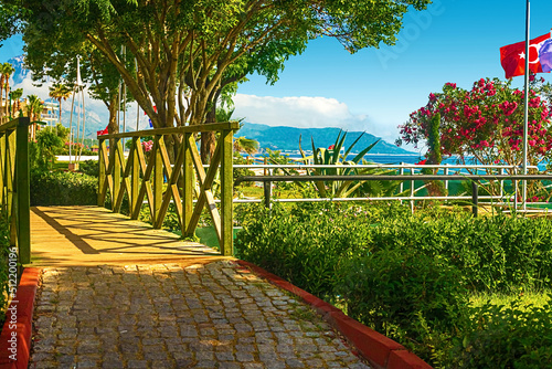 Fototapete View of the path and the bridge in a green garden with green trees along the embankment in Kemer, Turkey