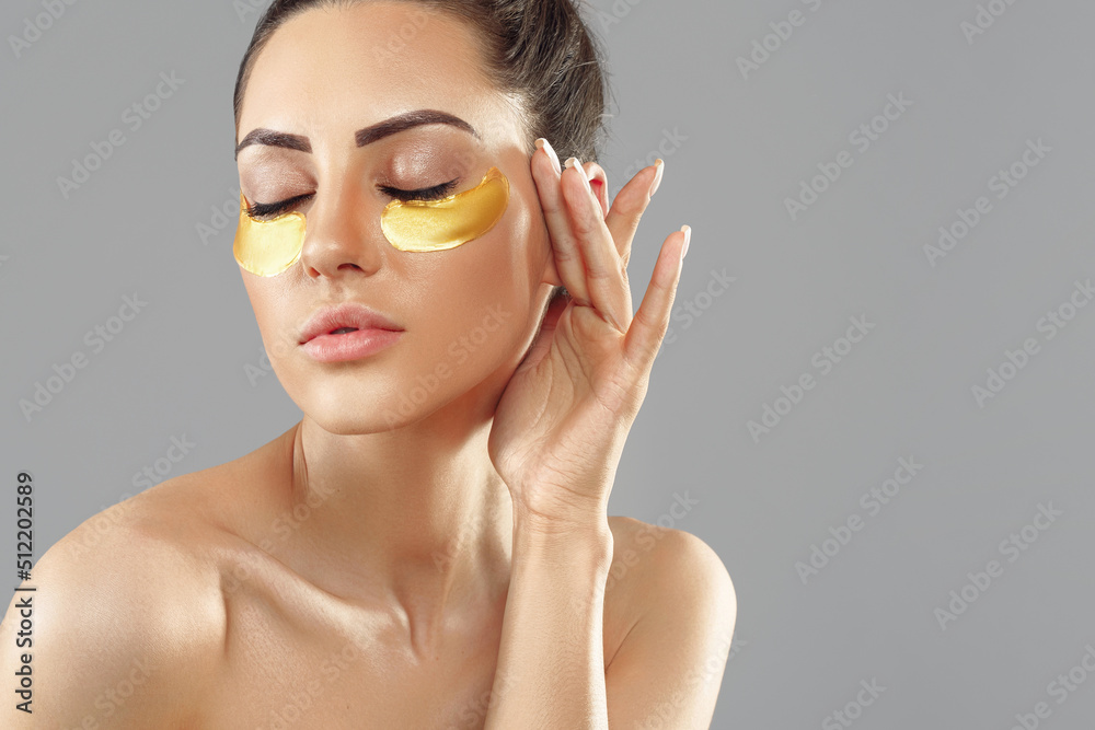 Beautiful woman with gold patches for eyes on a grey background. Patch under eyes. Collagen mask. Portrait of female, facial treatmen Beautiful woman with gold patches for eyes on a grey background. Patch under eyes. Collagen mask. Portrait of female, facial treatmen