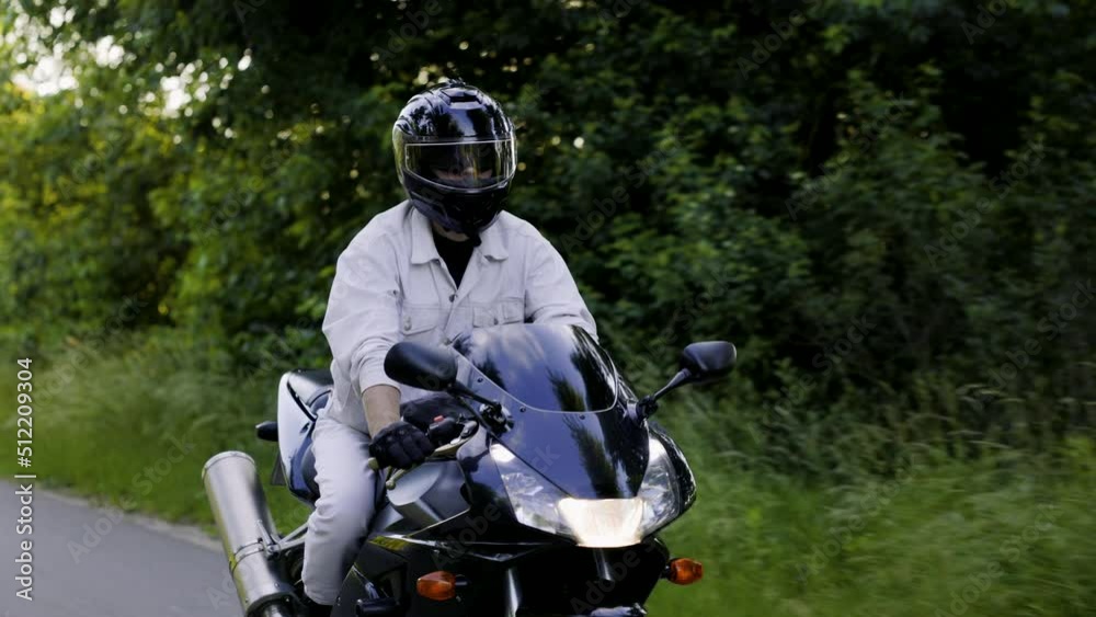 Man in helmet drives his sport black motorcycle on the summer road between trees and bushes during sunset. Motorcyclist dressed in a white denim suit. Medium shot Man in helmet drives his sport black motorcycle on the summer road between trees and bushes during sunset. Motorcyclist dressed in a white denim suit. Medium shot