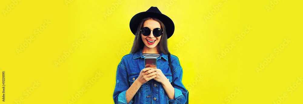 Obraz premium Portrait of stylish happy smiling young woman model with smartphone wearing black round hat, jean jacket on yellow background, blank copy space for advertising text