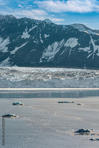 Disenchantment Bay, Alaska, USA - July 21, 2011: Portrait of floating ice in front of Hubbard glacier blue ice wall with tall mountain range and snow patches in back under blue cloudscape