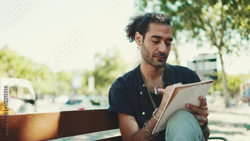 Young italian guy with ponytail and stubble sits on street bench and makes sketches with pen on sheet of paper on cityscape background