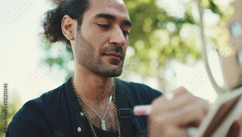 Close-up, young italian guy with ponytail and stubble sits on street bench and makes sketches with pen on sheet of paper on cityscape background