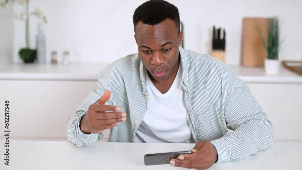 Young adult positive joyful successful african american man sitting at the table with a phone in his hands, rejoicing in victory, news or message, gesturing with hand, looking at the screen, smiling Young adult positive joyful successful african american man sitting at the table with a phone in his hands, rejoicing in victory, news or message, gesturing with hand, looking at the screen, smiling