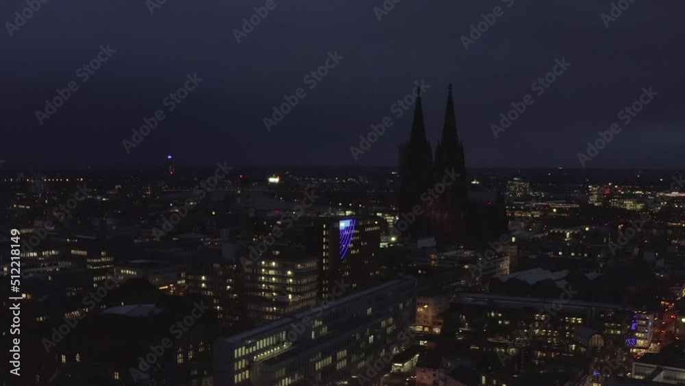 Forwards fly above night city. Buildings with lighted windows and illuminated streets in urban borough. Silhouette of Cologne Cathedral with tall towers. Cologne, Germany Forwards fly above night city. Buildings with lighted windows and illuminated streets in urban borough. Silhouette of Cologne Cathedral with tall towers. Cologne, Germany
