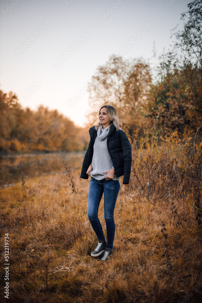 Shining blonde woman standing on grass near river, looking away smiling with black jacket on shoulders and hands on sweater with trees covered with sunset in background. Autumn walk in forest. Shining blonde woman standing on grass near river, looking away smiling with black jacket on shoulders and hands on sweater with trees covered with sunset in background. Autumn walk in forest.